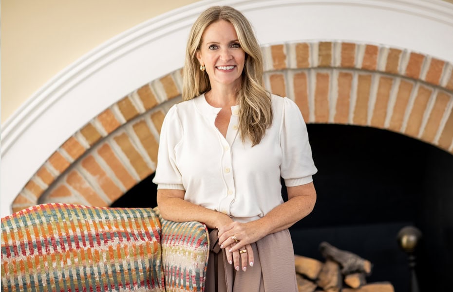 A woman with long blonde hair stands indoors in front of a brick fireplace, resting one hand on a colorful patterned blanket draped over a chair.