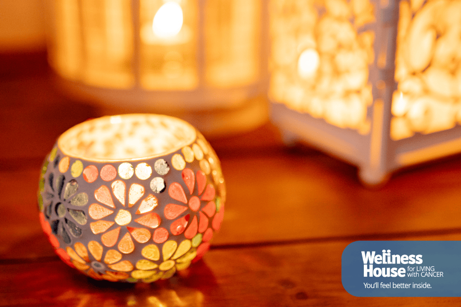 A colorful mosaic candle holder and two lanterns glow on a wooden surface, creating a warm and inviting atmosphere. The Wellness House logo appears in the bottom right corner, highlighting their support for those affected by prostate cancer.