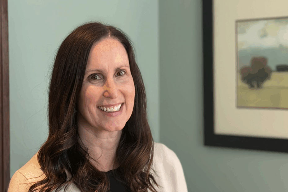 Portrait of Jamie Shifley, MS, RDN, LDN, a woman with long brown hair smiles at the camera, standing indoors in front of a teal wall and a framed landscape painting.