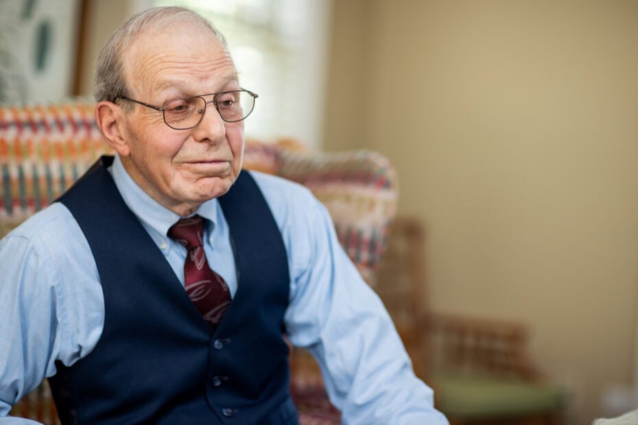 Elderly man with glasses wearing a blue shirt, tie, and vest sits on a patterned armchair in a softly lit room, reflecting on the benefits of cancer wellness programs.