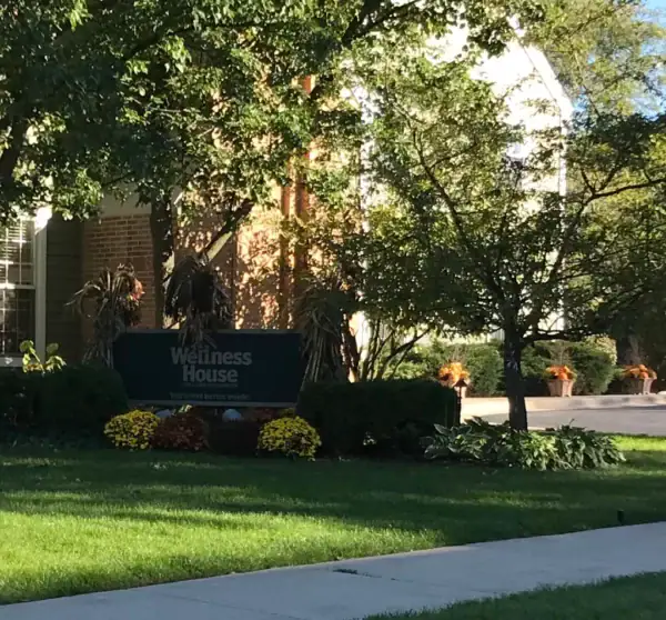 A green sign reading “Wellness House” stands among bushes and flowers in front of a brick building, surrounded by trees and a well-kept lawn.