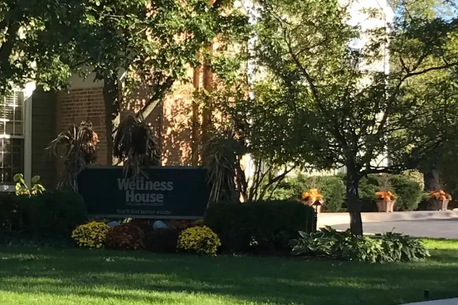 A green sign reading “Wellness House” stands among bushes and flowers in front of a brick building, surrounded by trees and a well-kept lawn.