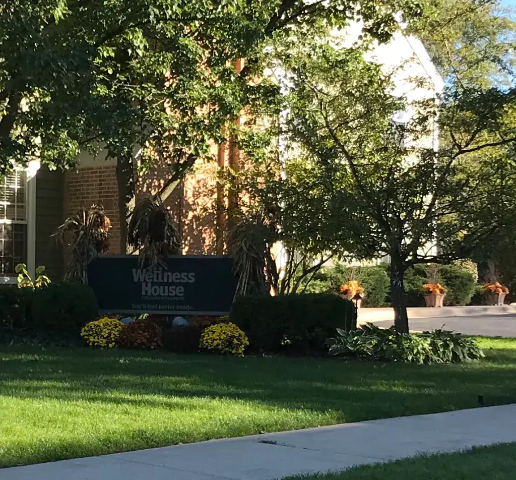 A green sign reading “Wellness House” stands among bushes and flowers in front of a brick building, surrounded by trees and a well-kept lawn.