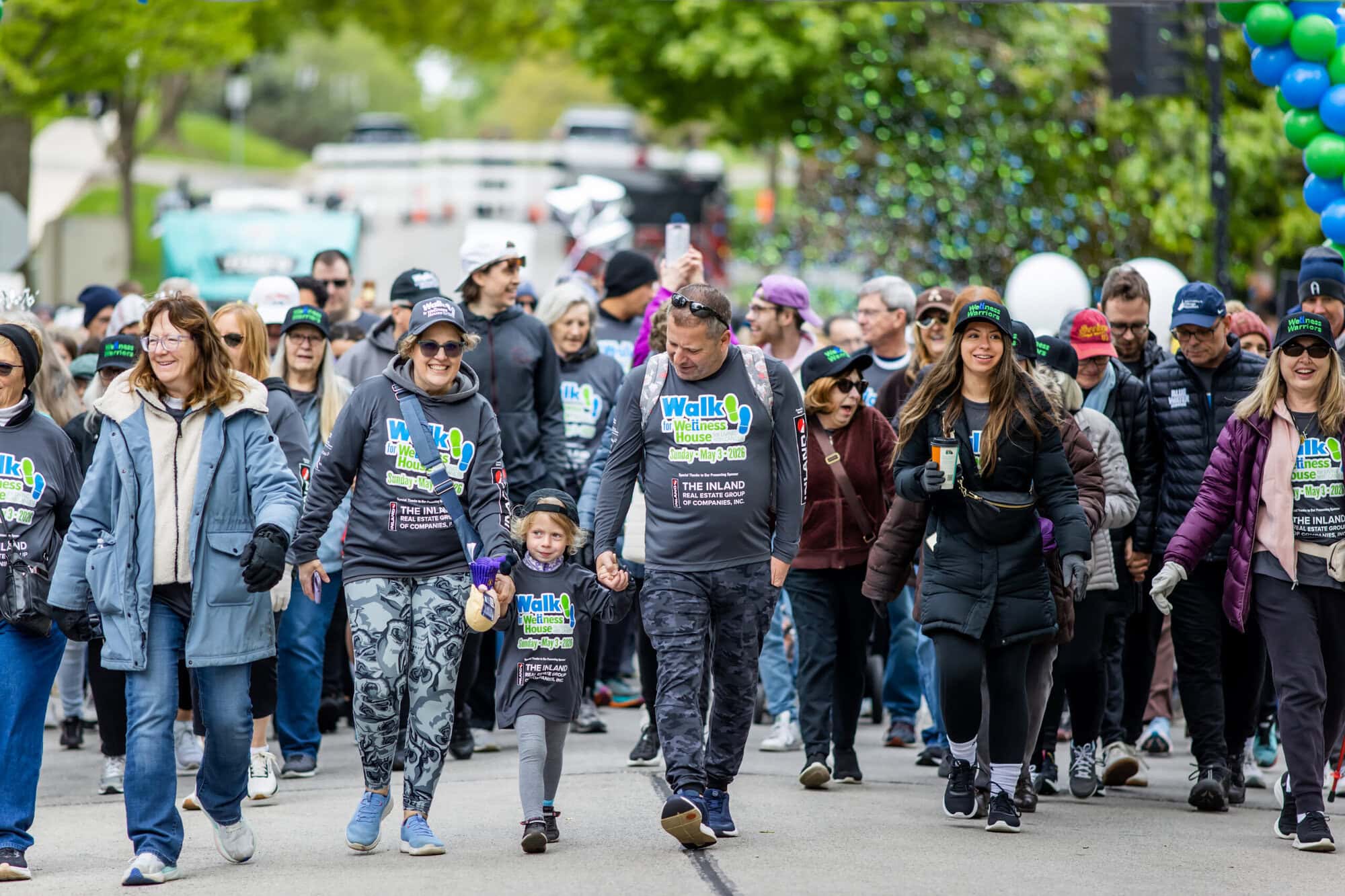 A large group of people wearing "Walk" event shirts and casual clothes walk together outdoors on a street, surrounded by balloons, trees, and the lively atmosphere of community events.