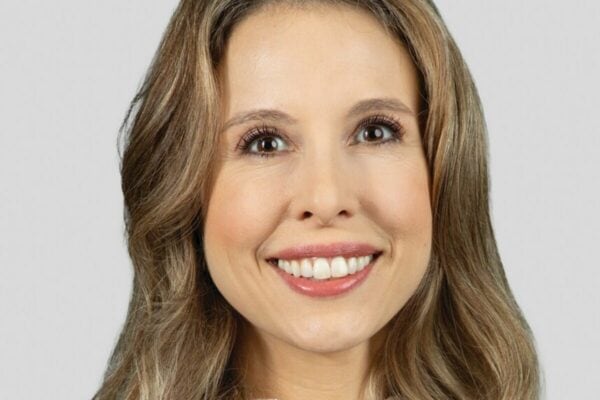 A woman with long, wavy light brown hair smiles at the camera against a light gray background.