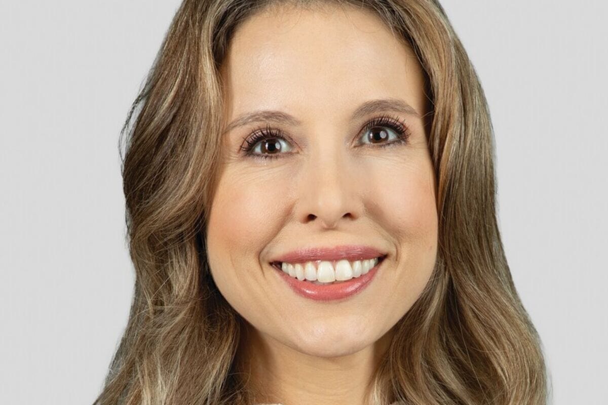 A woman with long, wavy light brown hair smiles at the camera against a light gray background.