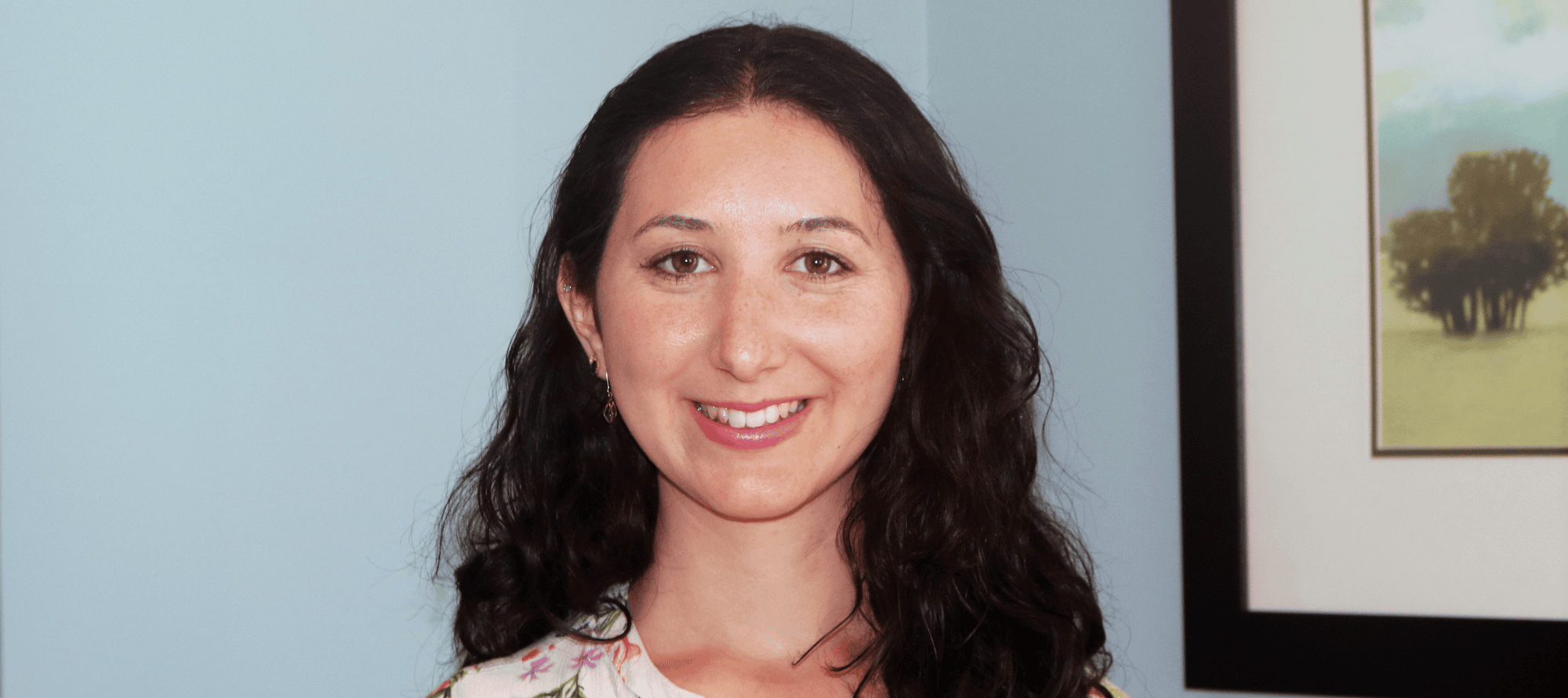 A woman with long, dark, wavy hair smiles at the camera, standing in front of a light blue wall and a framed picture—radiating the confidence often seen in cancer wellness experts.
