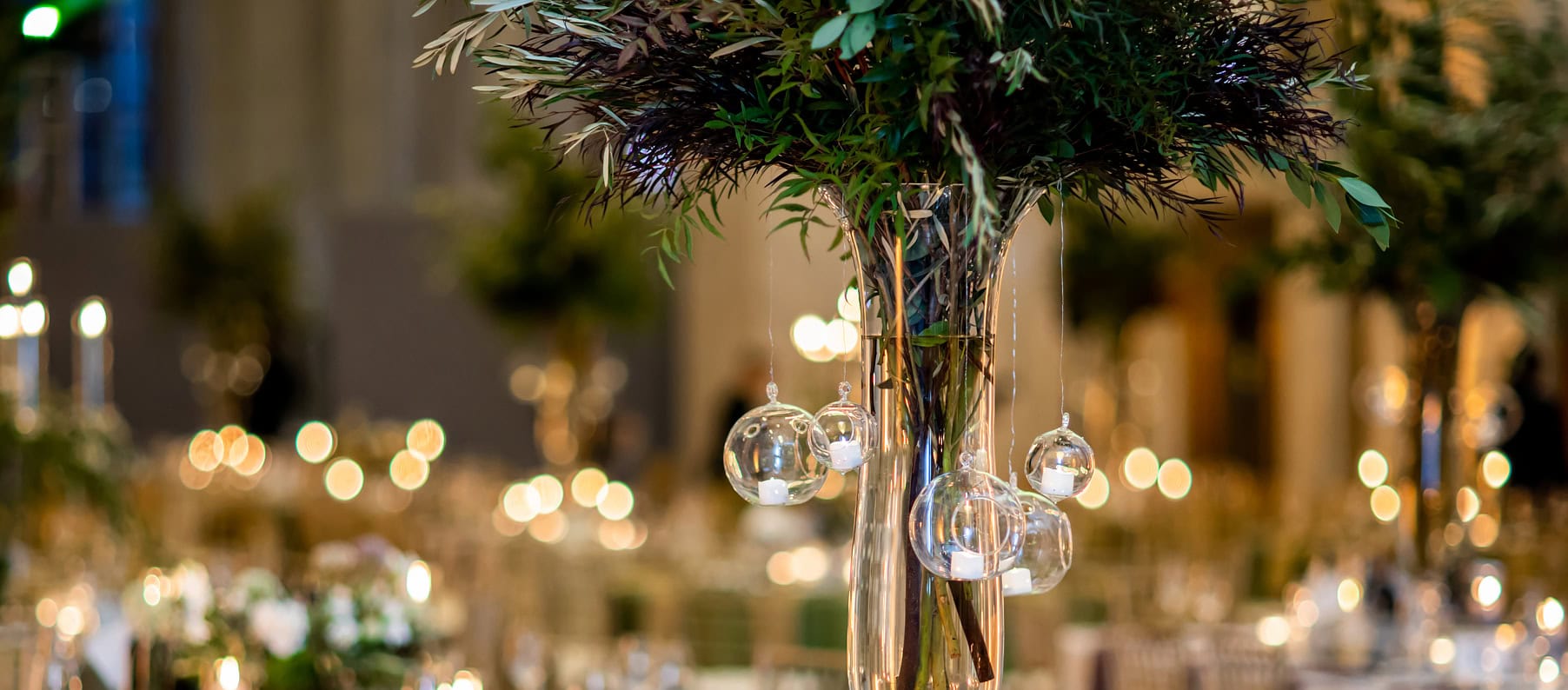 Tall floral centerpiece with green foliage and hanging glass ornaments on a table in an elegantly decorated venue with blurred lights in the background.
