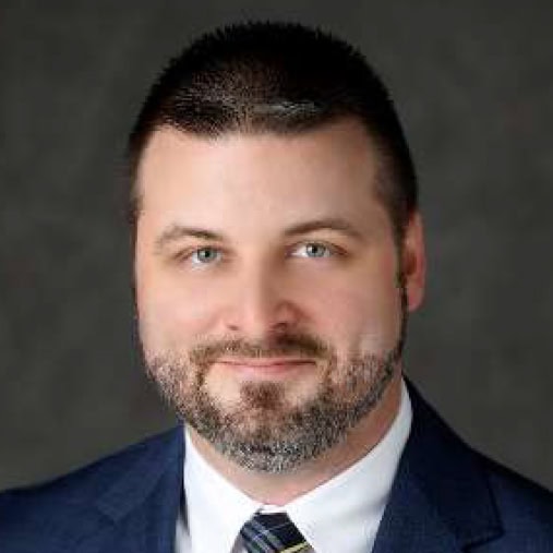 Man with short dark hair and beard wearing a dark suit, white shirt, and striped tie, posing against a plain dark background.