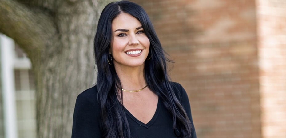 A woman with long black hair, wearing a black V-neck top and gold necklace, stands outdoors in front of a tree and brick wall, smiling at the camera—radiating the warmth and confidence of someone who could be among today’s leading cancer wellness experts.