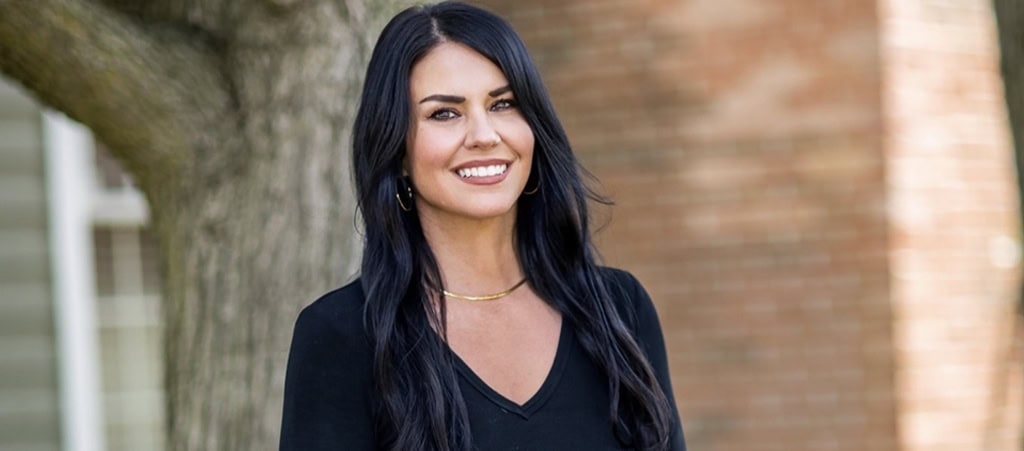 A woman with long black hair, wearing a black V-neck top and gold necklace, stands outdoors in front of a tree and brick wall, smiling at the camera—radiating the warmth and confidence of someone who could be among today’s leading cancer wellness experts.