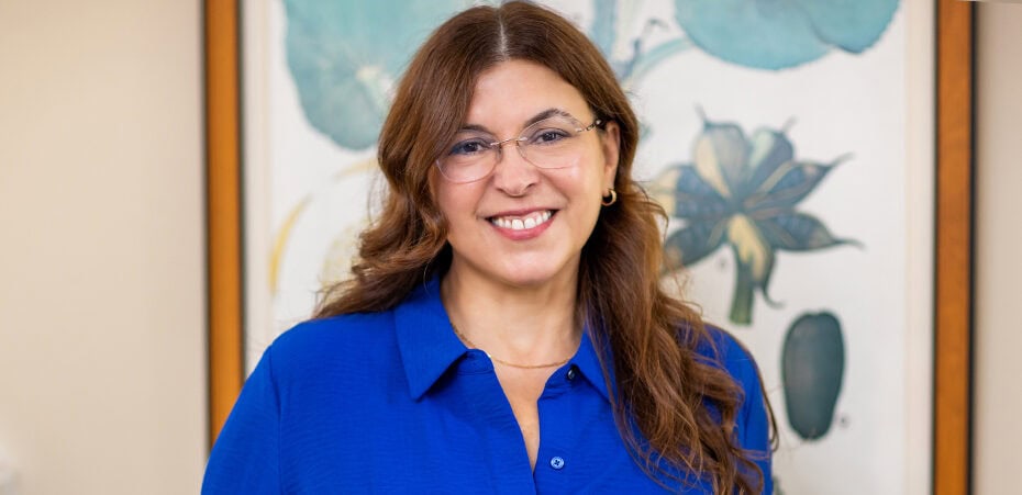 Woman with long brown hair and glasses, wearing a blue shirt, standing indoors in front of a framed botanical artwork, smiling at the camera—embodying the warmth and approachability often associated with cancer wellness experts.