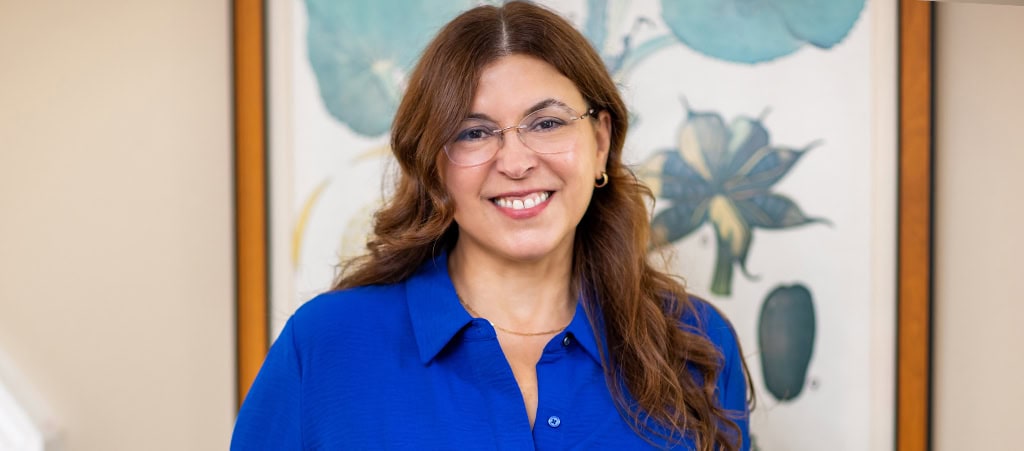 Woman with long brown hair and glasses, wearing a blue shirt, standing indoors in front of a framed botanical artwork, smiling at the camera—embodying the warmth and approachability often associated with cancer wellness experts.
