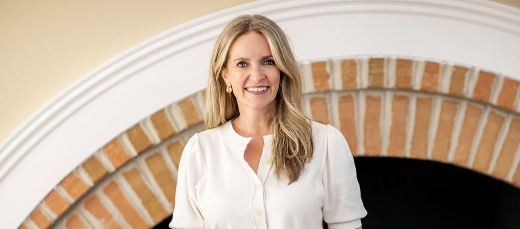 Woman with long blonde hair wearing a white blouse stands in front of an arched brick fireplace, smiling at the camera—radiating the warmth and confidence often found in cancer wellness experts.