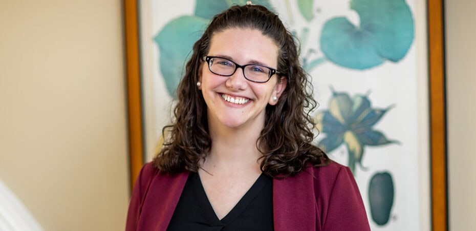 A person with long curly hair and glasses, wearing a maroon blazer and black shirt, smiles in front of a framed floral artwork, embodying the approachable professionalism often seen among cancer wellness experts.