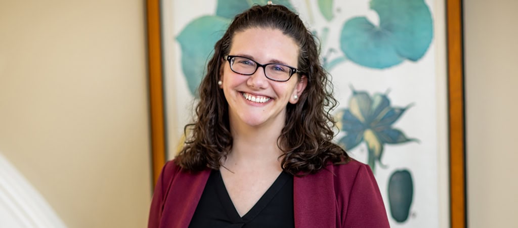 A person with long curly hair and glasses, wearing a maroon blazer and black shirt, smiles in front of a framed floral artwork, embodying the approachable professionalism often seen among cancer wellness experts.