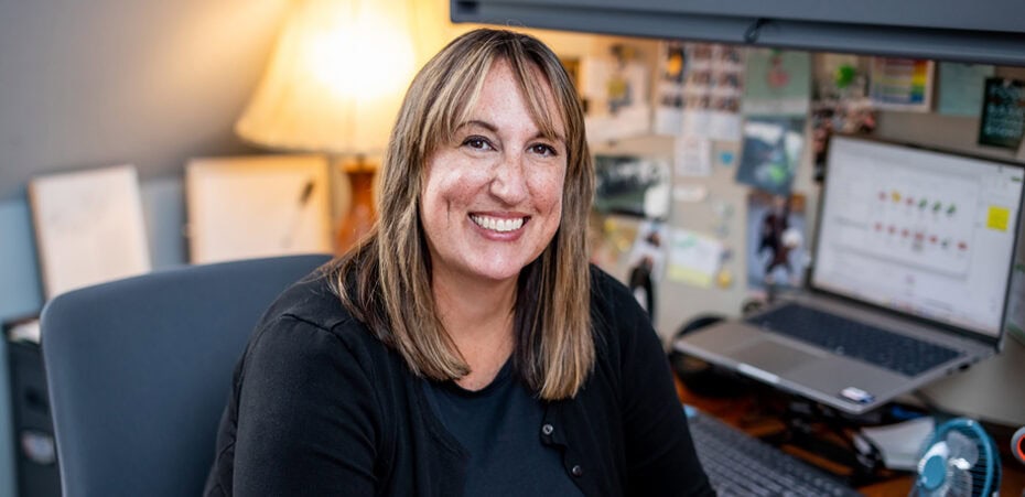 Woman with straight, shoulder-length hair smiles while sitting at a desk with a laptop, lamp, and bulletin board in a home office setting, embodying the warmth and professionalism of cancer wellness experts.