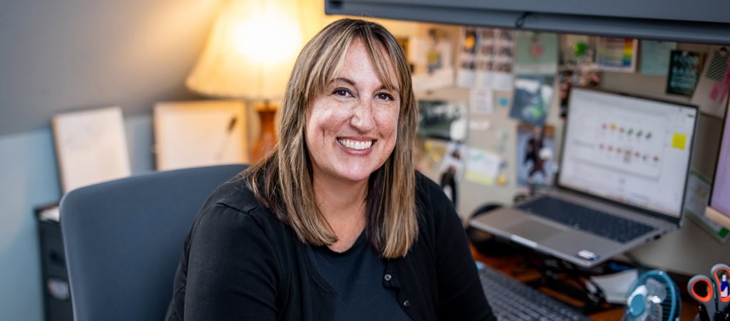 Woman with straight, shoulder-length hair smiles while sitting at a desk with a laptop, lamp, and bulletin board in a home office setting, embodying the warmth and professionalism of cancer wellness experts.