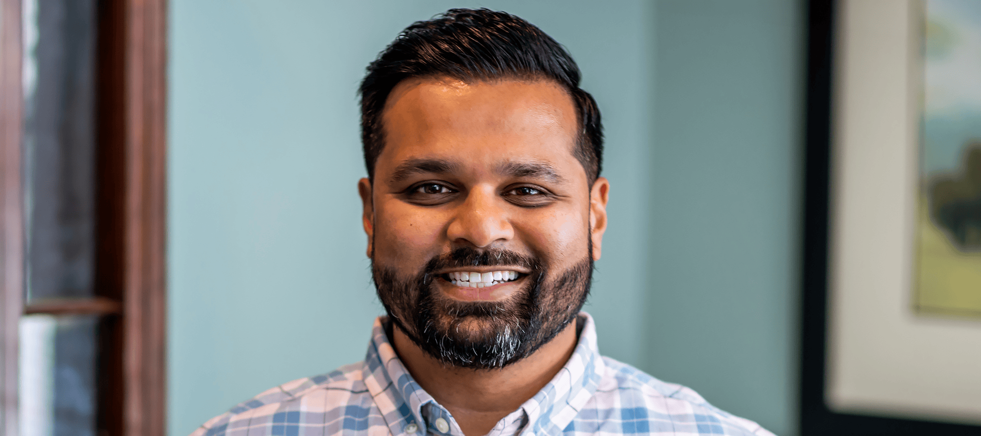 A man with dark hair and a beard smiles at the camera, wearing a light blue and white checked shirt, standing indoors with a blurred background, embodying the warmth often associated with cancer wellness experts.
