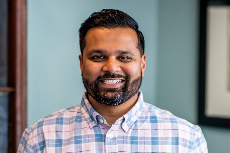A man with short dark hair and a beard, wearing a light plaid button-up shirt, smiles while standing indoors in front of a blue wall.