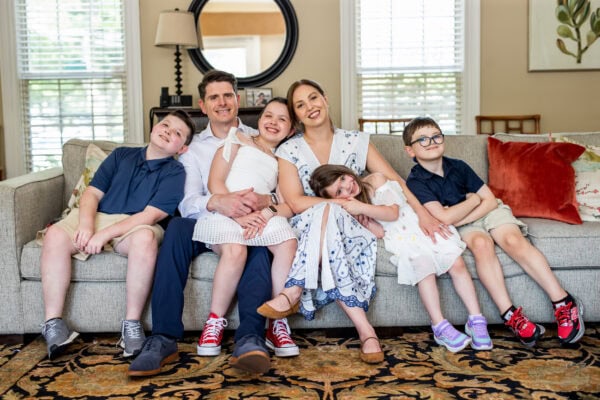 A family of two adults and four children sit together on a couch in a living room, all smiling at the camera.