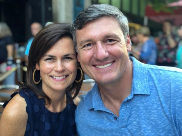 A woman and a man are smiling and posing closely together outdoors, with other people blurred in the background.