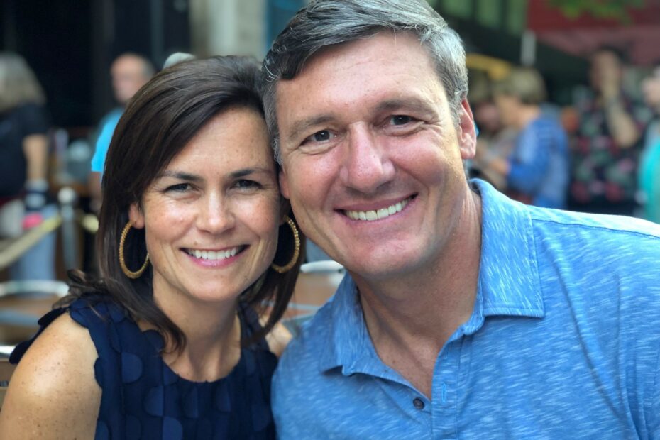 A woman and a man are smiling and posing closely together outdoors, with other people blurred in the background.