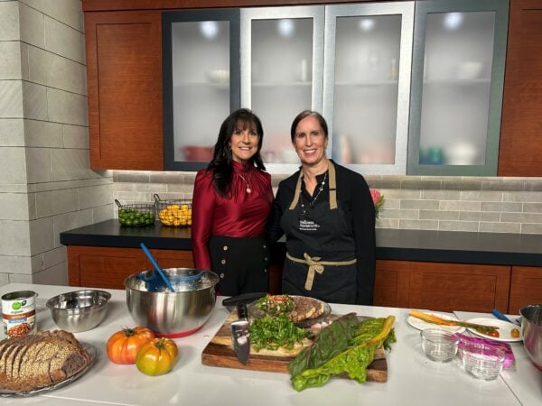 Two women stand in a kitchen behind a counter with ingredients and prepared food for a cooking segment.