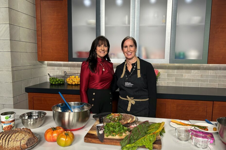 Two women stand in a kitchen behind a counter with ingredients and prepared food for a cooking segment.