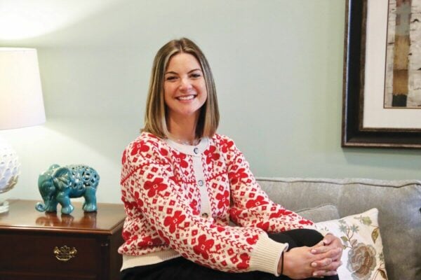 A woman smiling while wearing a red and white patterned cardigan, sitting on a sofa.