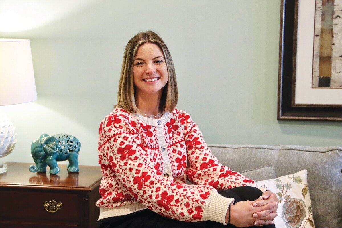 A woman smiling while wearing a red and white patterned cardigan, sitting on a sofa.